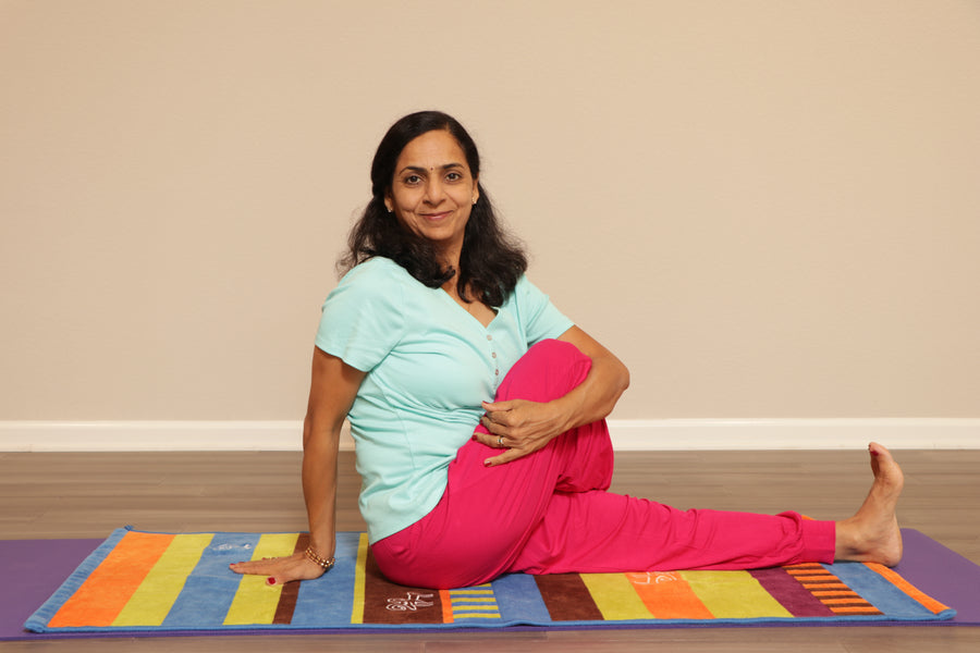 A woman practicing yoga on a colorful mat, sitting with her left hand on the floor, her right arm raised, wearing a light blue top and pink pants.